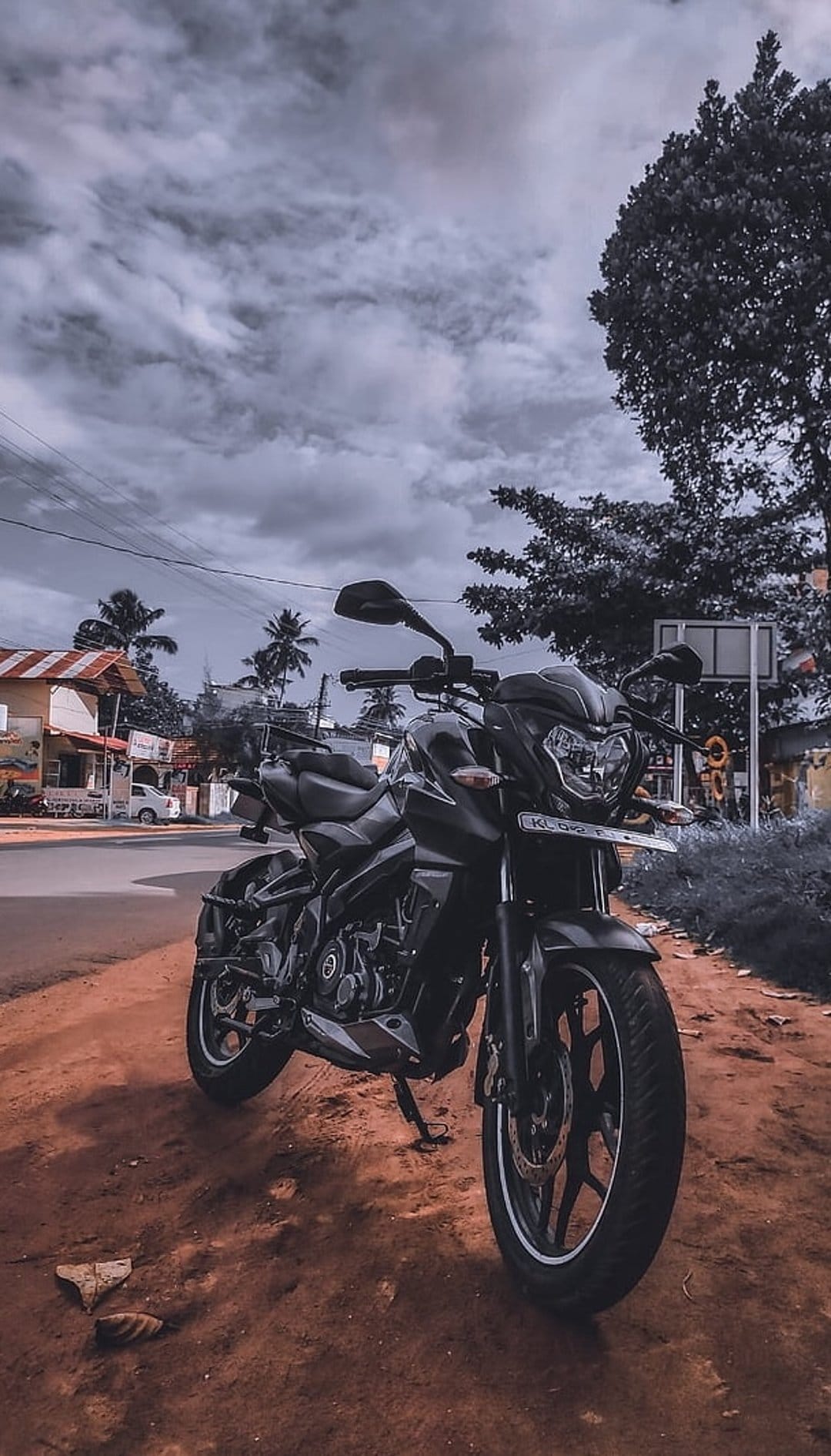 Black motorcycle parked on a red dirt road under dark stormy clouds with palm trees in the background.