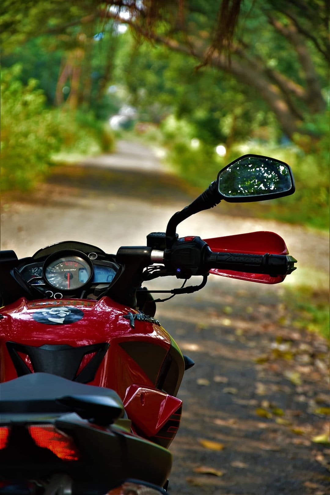Red motorcycle handlebar and rearview mirror reflecting a dirt road surrounded by dense green forest trees.