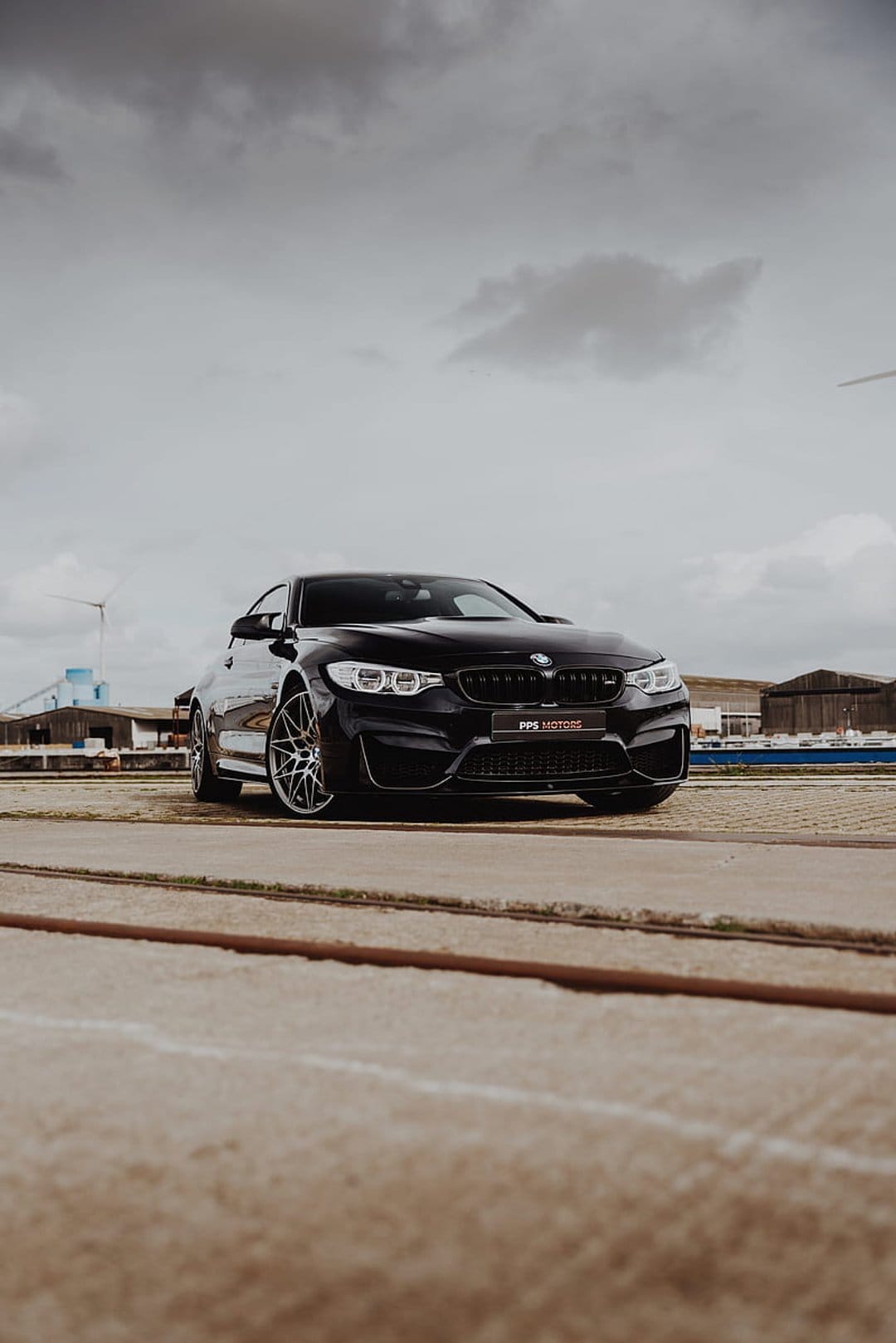 Black BMW M4 performance coupe on concrete near train tracks under a cloudy industrial sky with wind turbines.