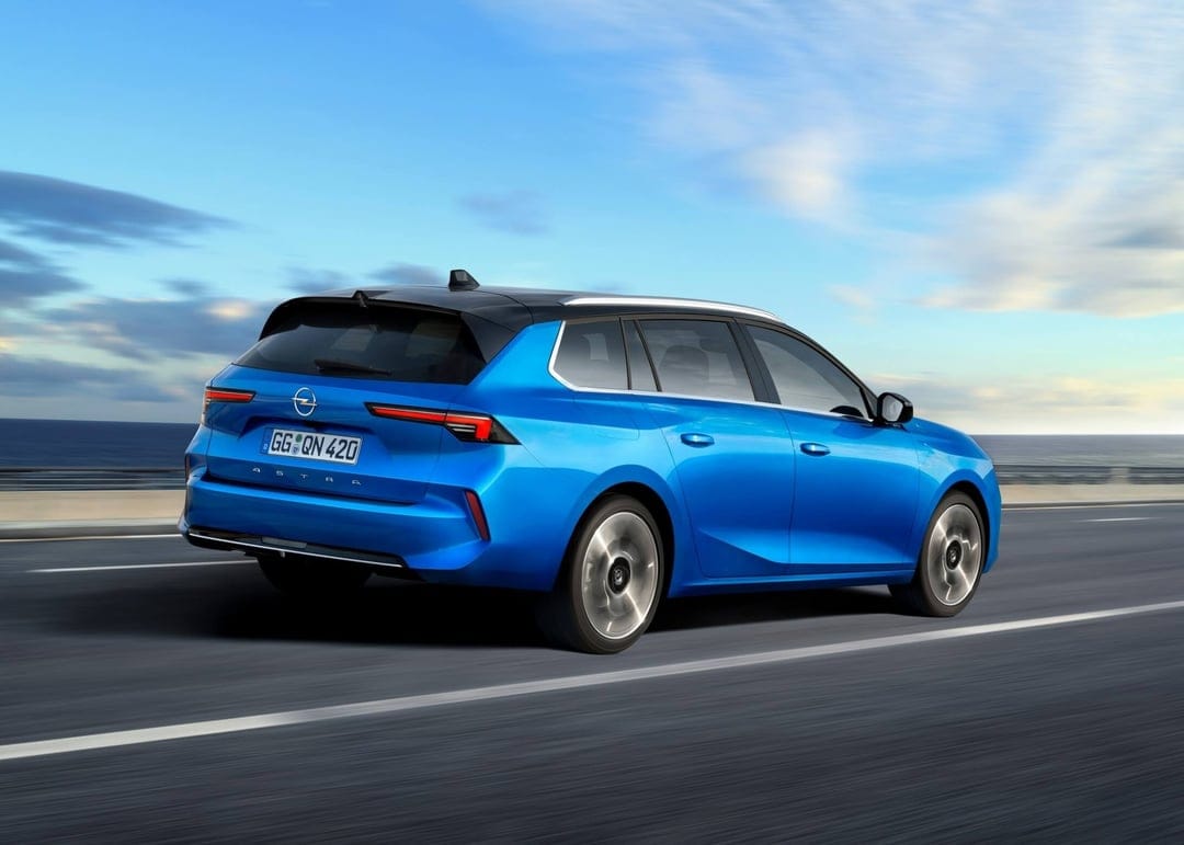 Blue station wagon driving on a coastal highway under a bright blue sky with motion blur on the road surface.