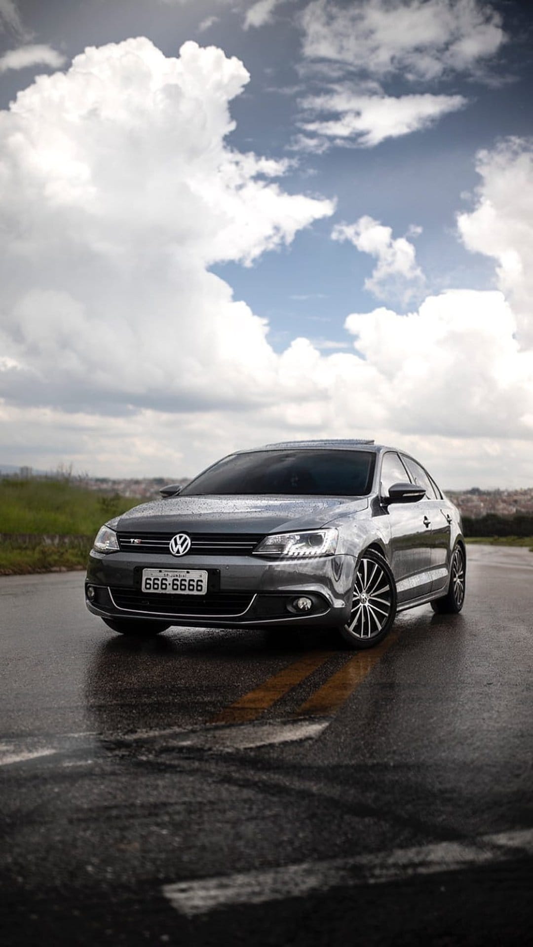 Gray Volkswagen Jetta sedan with glowing headlights on a wet asphalt road reflecting a dramatic cloudy sky.