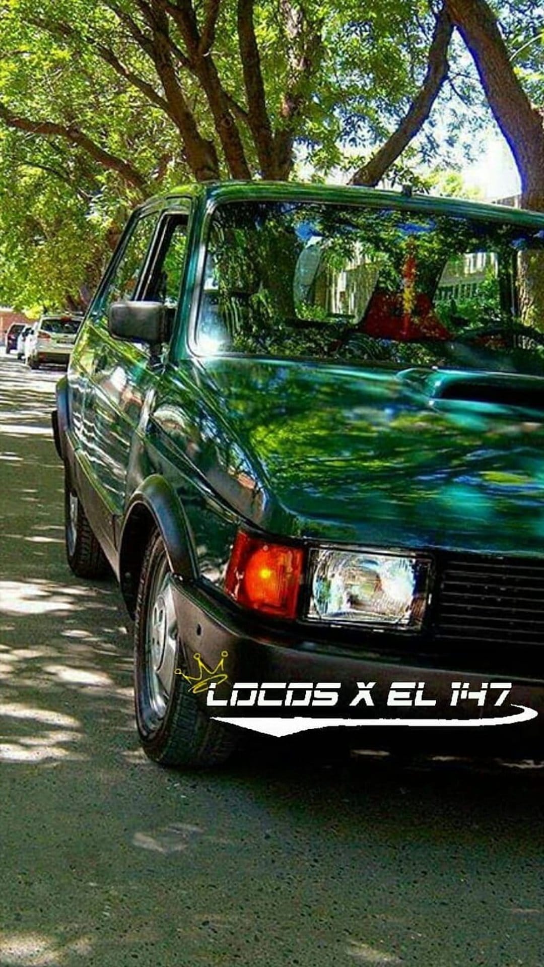 Green Fiat 147 parked on a street under dense tree foliage with dappled sunlight hitting the polished car body.