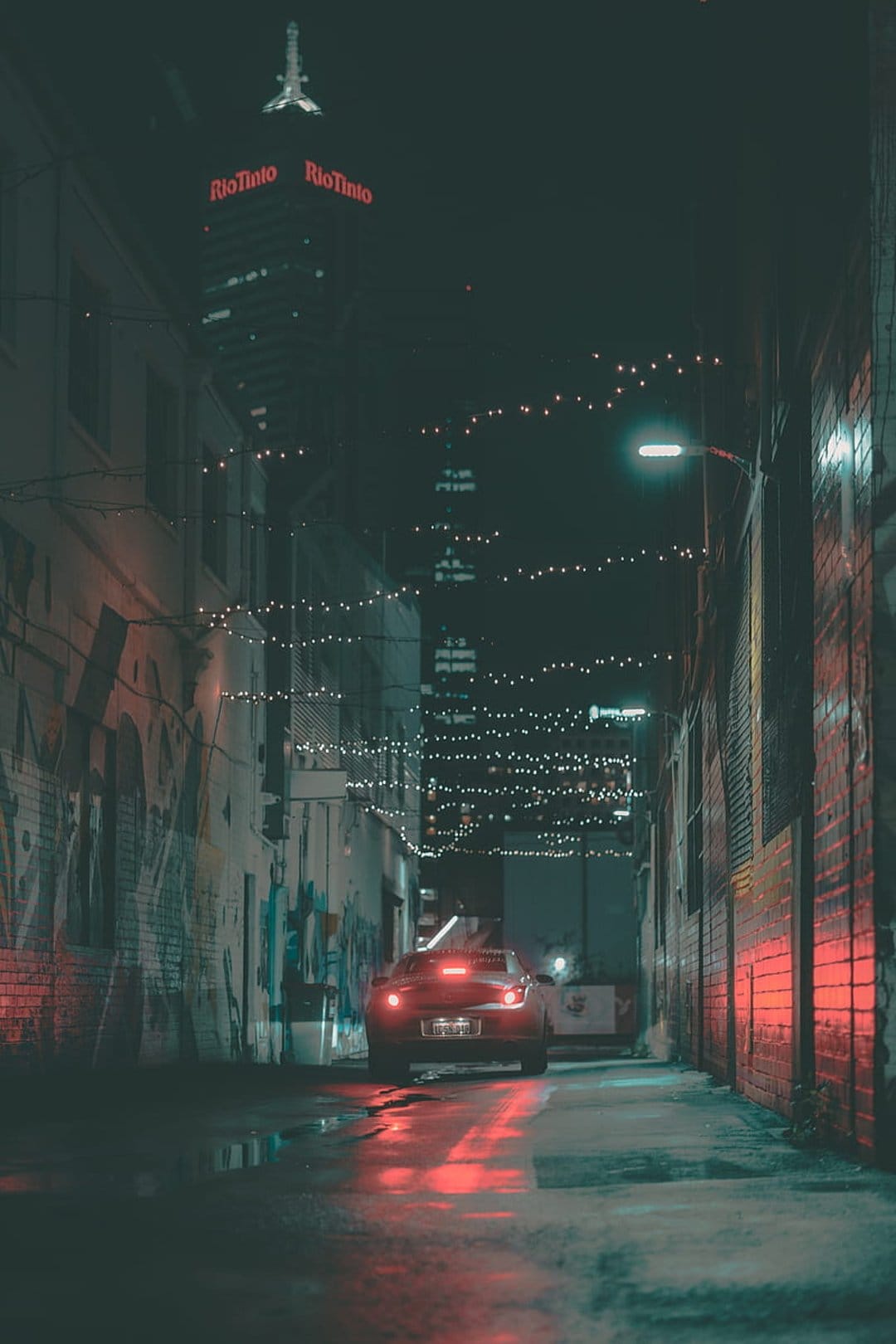 Red car taillights reflect on wet pavement in a dark alley with graffiti and a distant lit skyscraper.