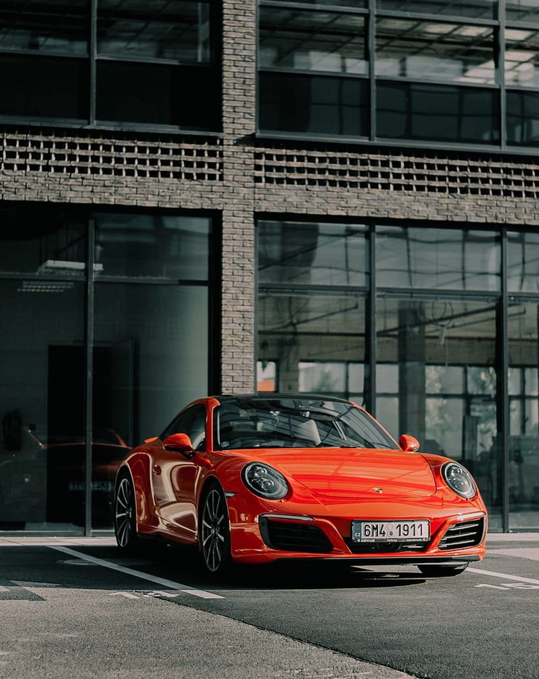 Orange Porsche 911 parked in front of a modern brick building with large glass windows and black roofing.