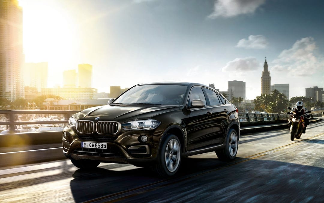 Dark brown BMW X6 SUV driving on a coastal city bridge during a bright sunset with a distant skyline.