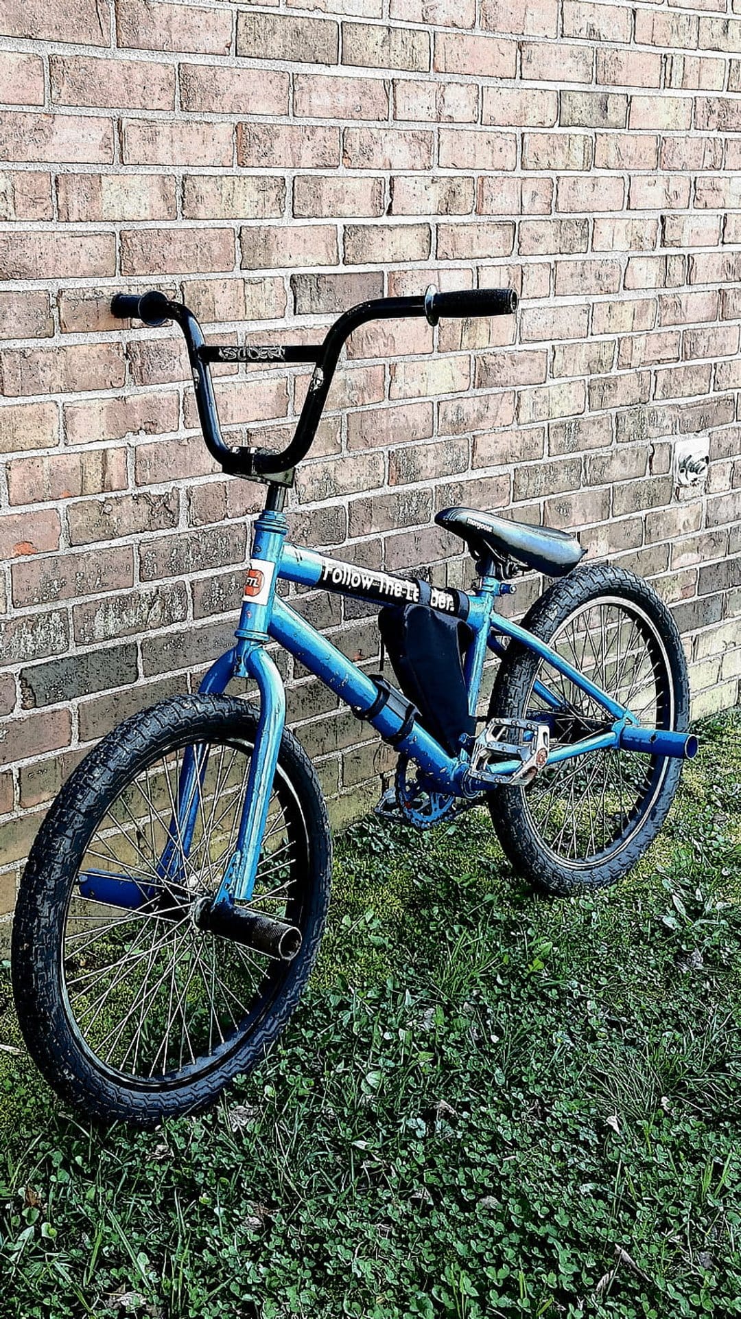 Blue BMX bike with black handlebars leans against a red brick wall on a patch of green clover and grass.