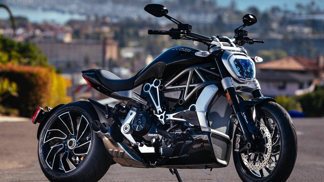 Black Ducati XDiavel S motorcycle with exposed frame and wide tires on a sunny road with blurred coastal background.