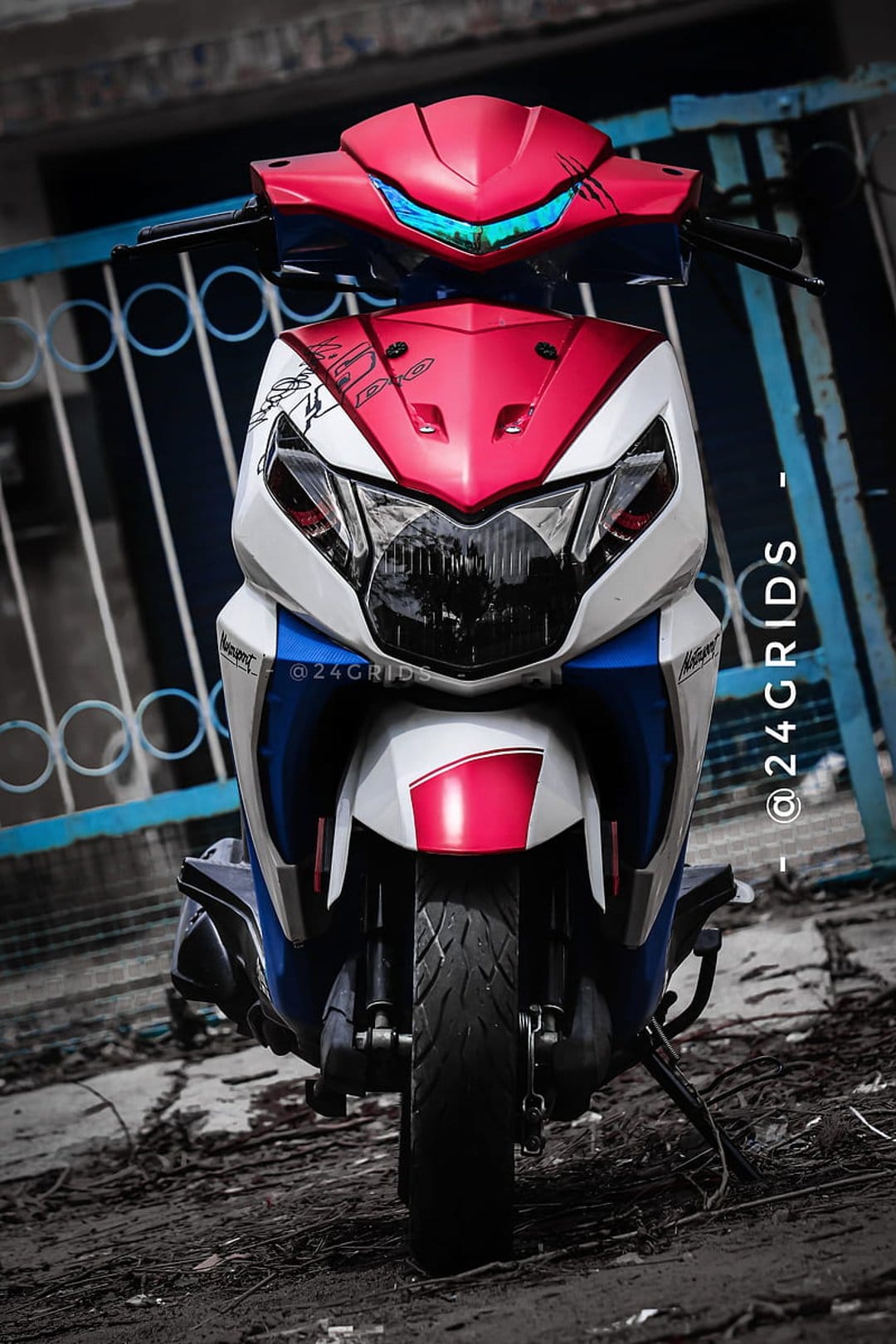 Front fairing of a red white and blue custom scooter with a modern headlight against a blue metal fence background.