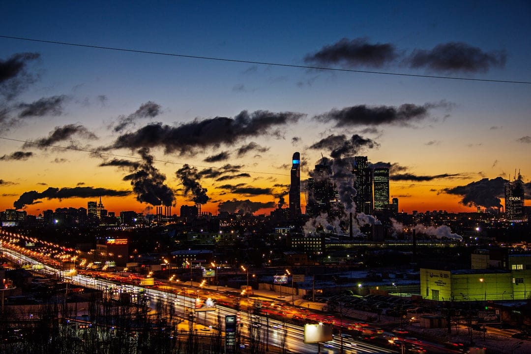 Towering factory chimneys release smoke plumes against a vivid orange and blue sunset sky above a glowing highway.