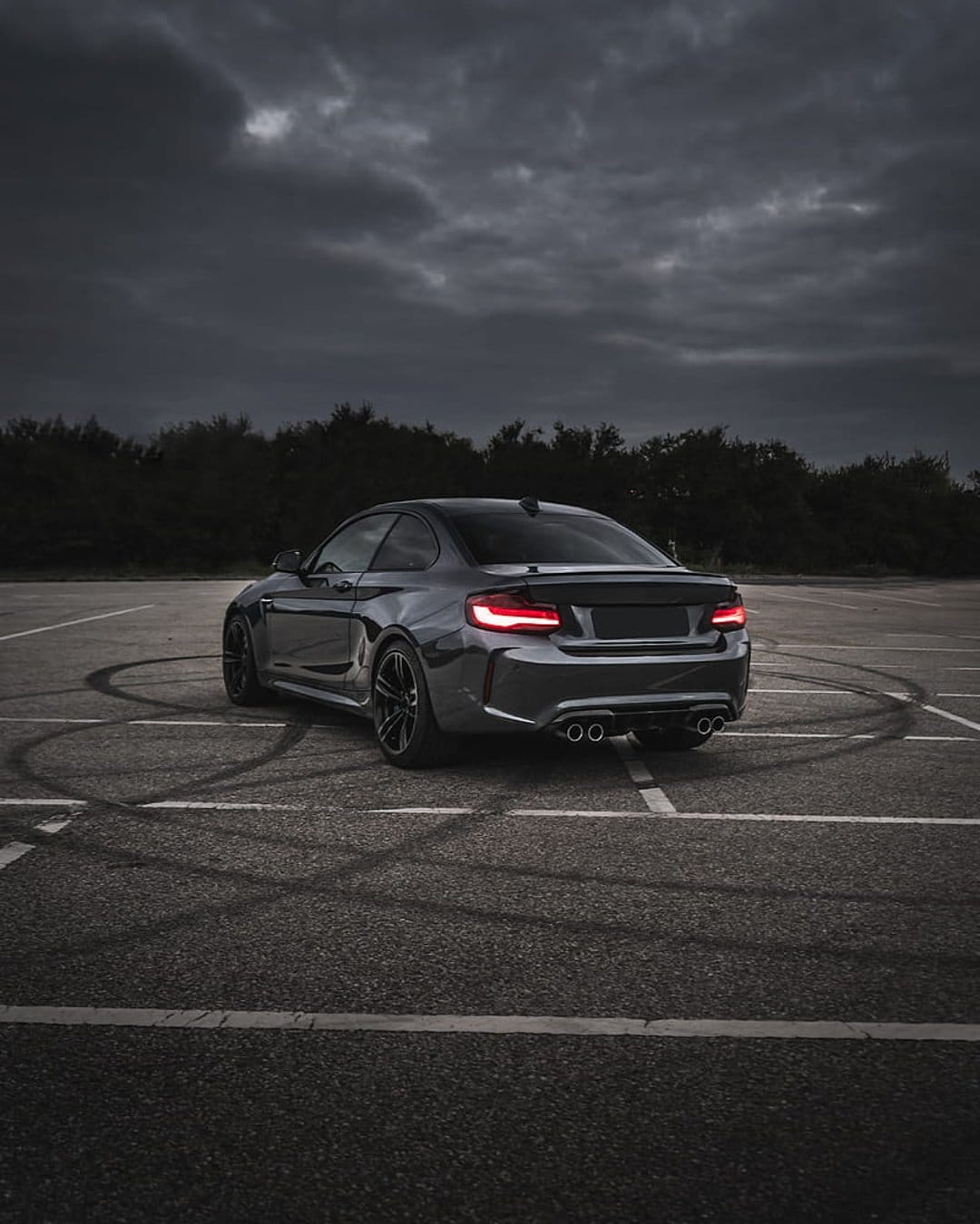 BMW M2 coupe with glowing red taillights and tire marks on asphalt under a dark, dramatic stormy sky.