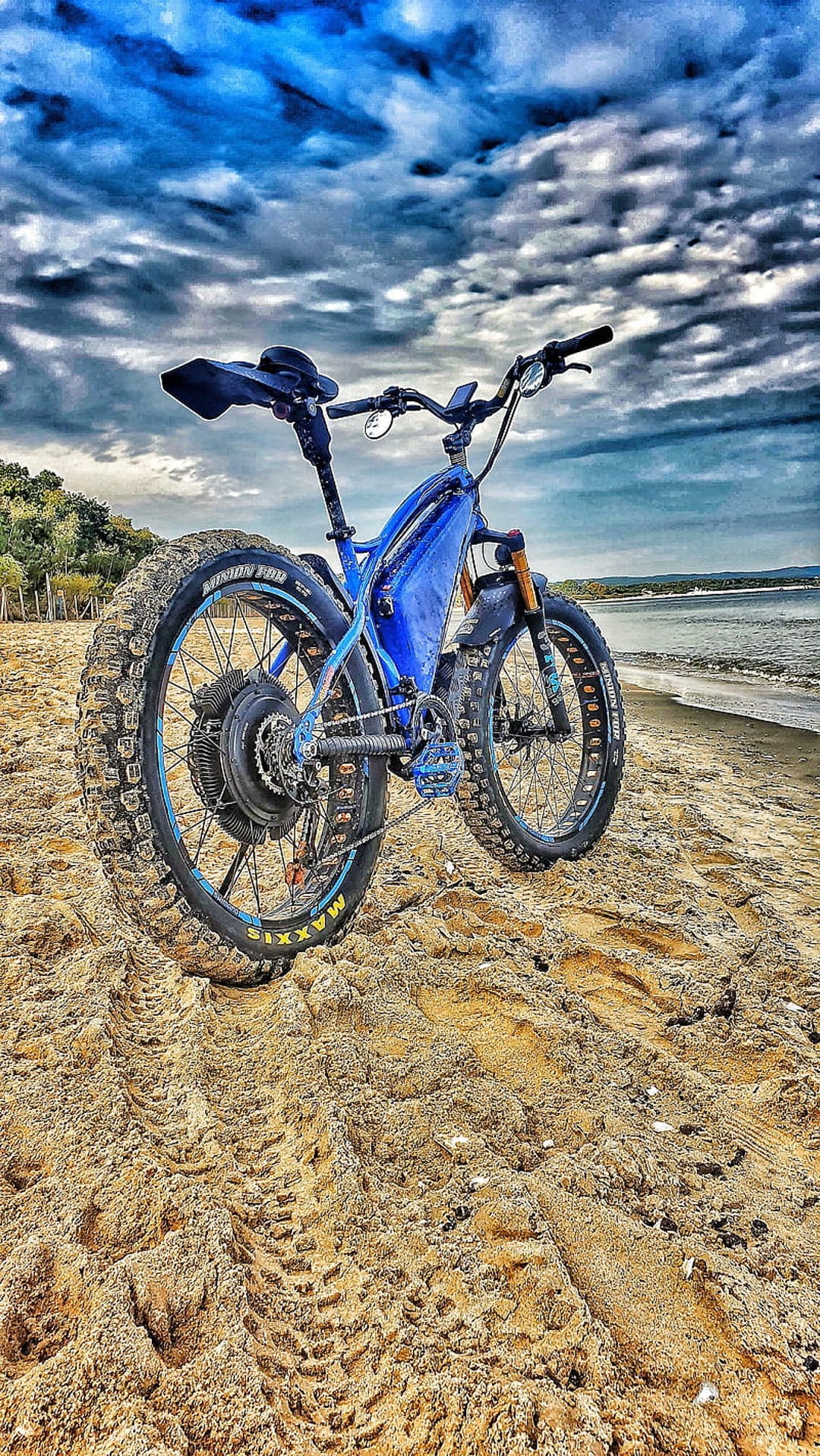 Blue electric fat tire bike parked on a sandy beach with ocean waves and cloudy sky in the background.