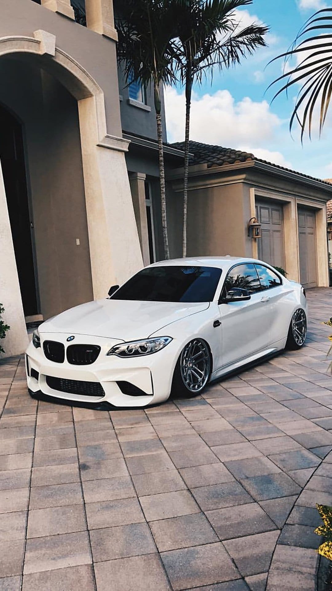 White BMW M2 with chrome wheels parked on cobblestones in front of a modern house under a bright blue sky.