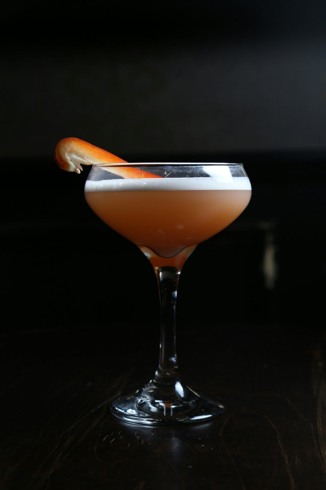 Amber cocktail in a coupe glass with citrus garnish and foam against a dark background for mobile screens.