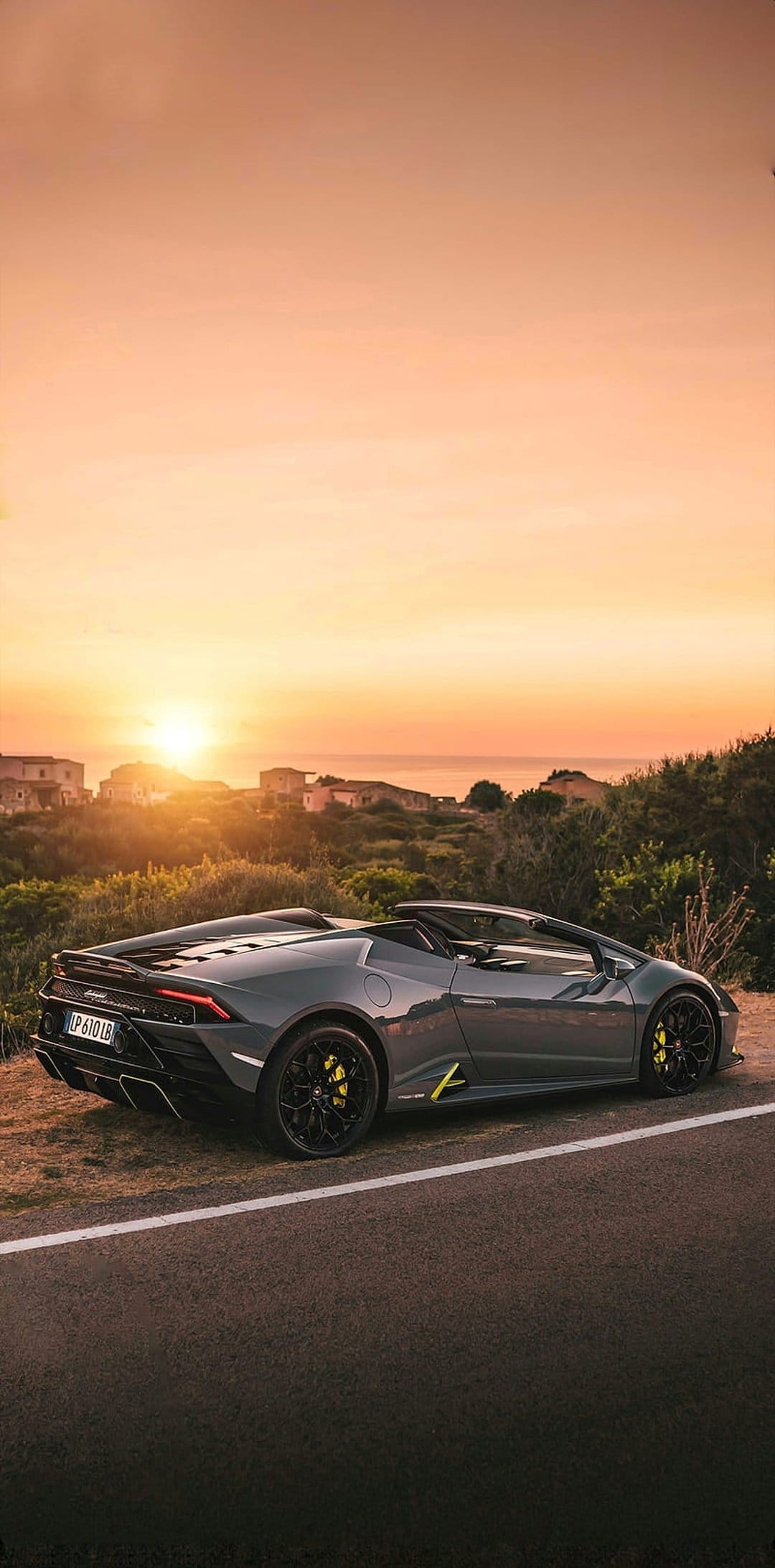 Gray Lamborghini Huracan Spyder on a coastal road during sunset with Mediterranean villas and ocean behind it.