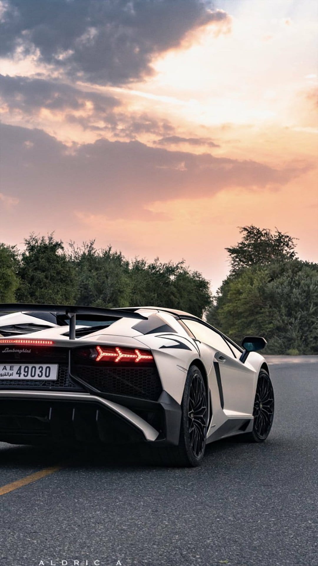 White Lamborghini Aventador rear with glowing LED tail lights parked against a dramatic orange sunset sky.
