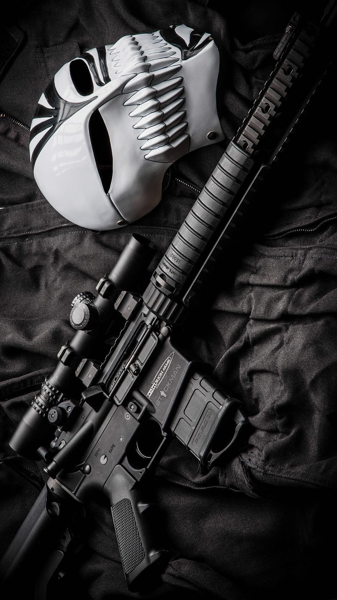 White skull mask with stylized teeth next to a black scoped rifle on dark textured fabric in high contrast.
