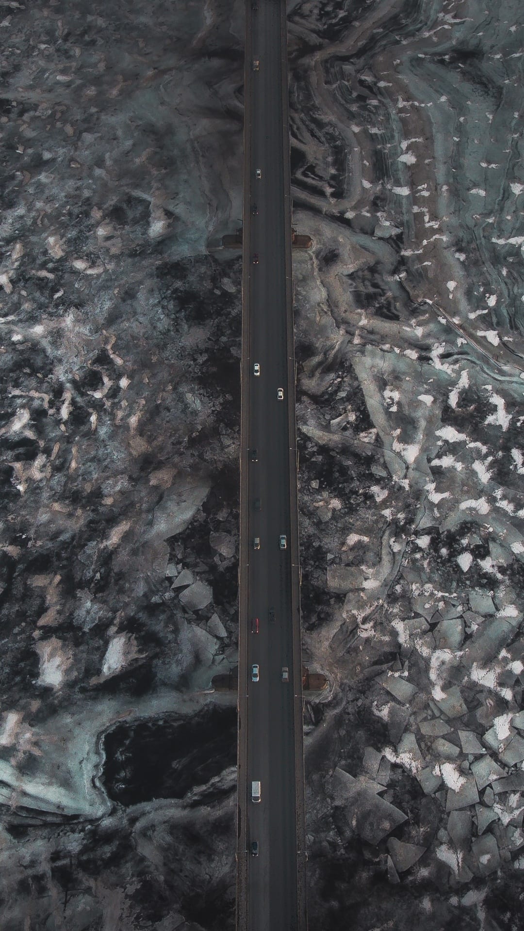 A narrow bridge crosses deep blue water covered in fractured white ice sheets from an aerial perspective.