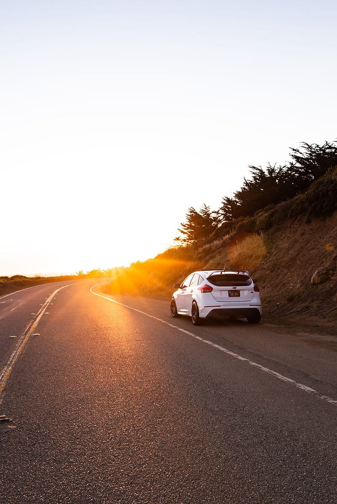 White car driving on a winding coastal road with a bright orange sunset over the ocean and green hills.