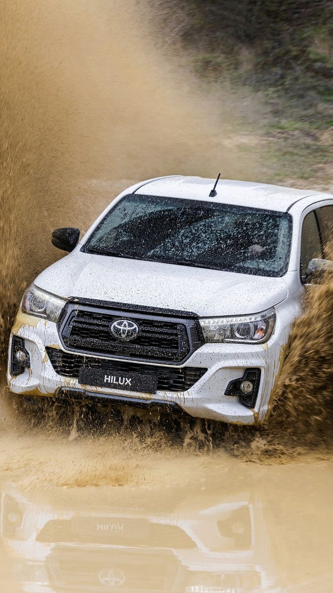 White Toyota Hilux truck splashes through a deep muddy puddle with water spraying from the rugged front tires.