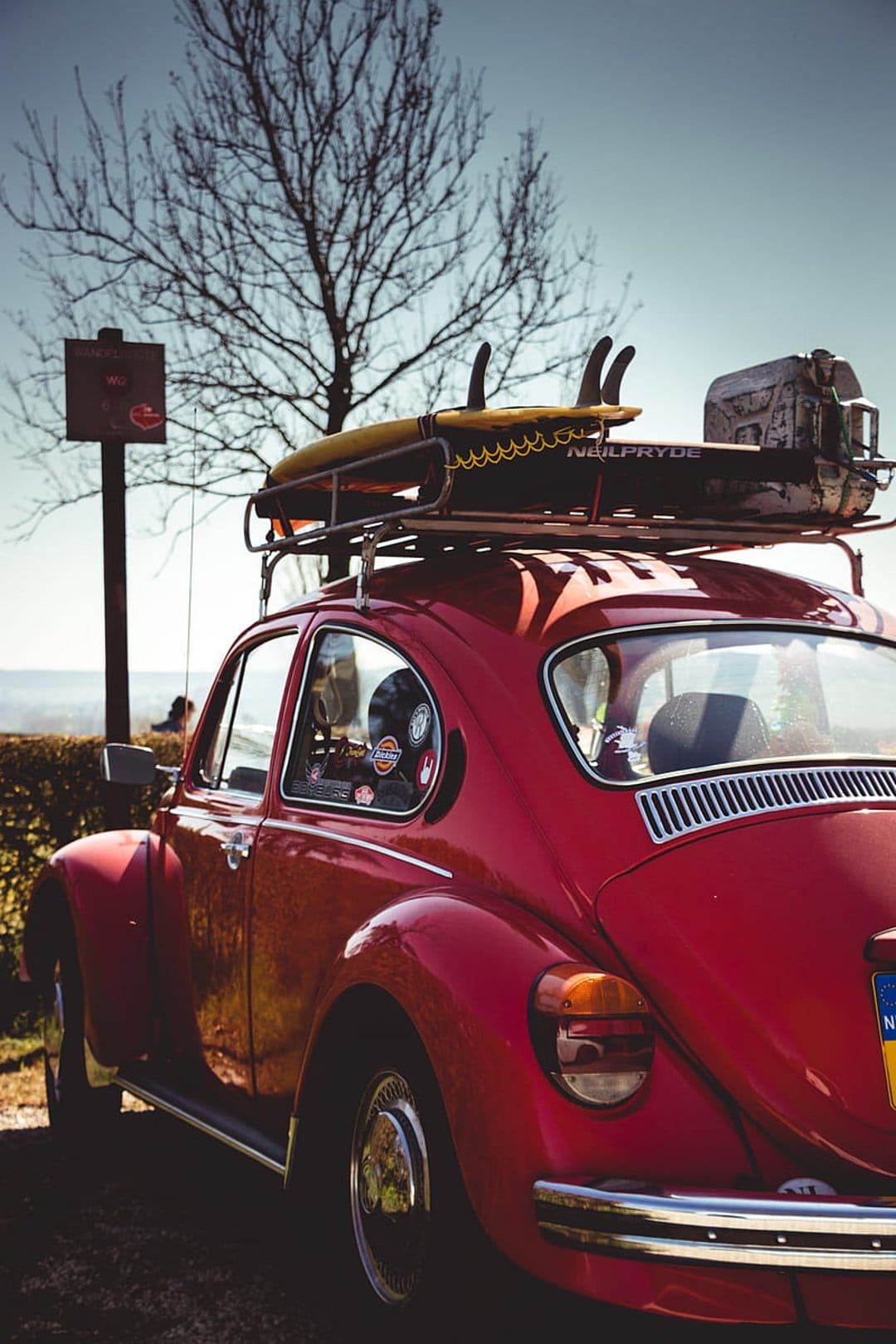 Red vintage Volkswagen Beetle with surfboards on a roof rack parked under a clear blue sky and a bare tree.