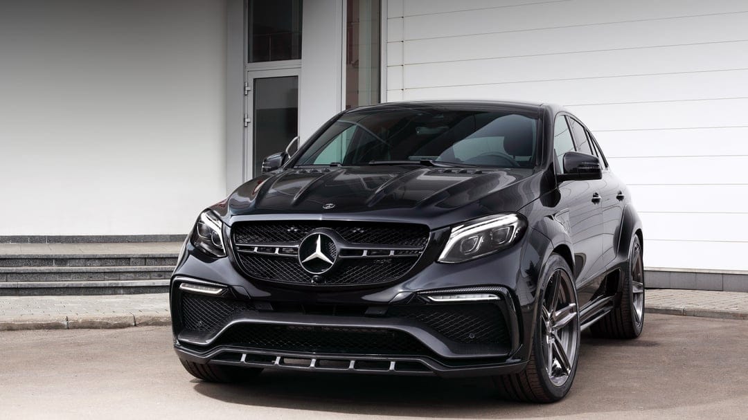Black Mercedes-Benz GLE Coupe with widebody kit and carbon fiber accents parked before a white building.