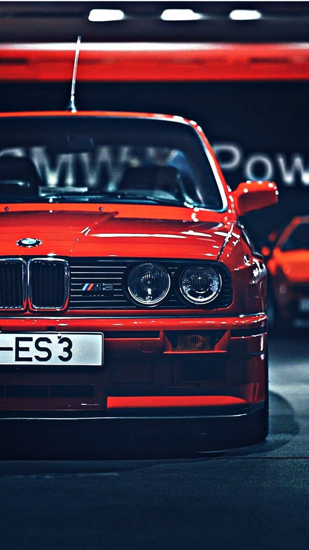 Red BMW M3 E30 front view featuring dual round headlights, black kidney grille, and vibrant paint in a garage.