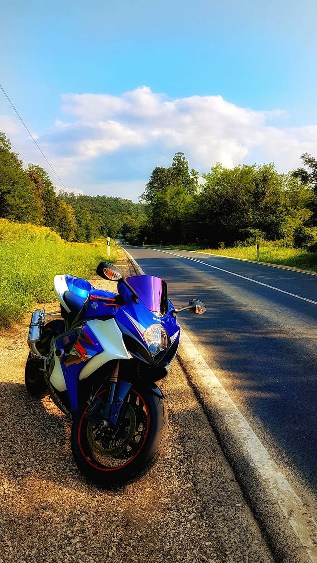 Blue and white sport motorcycle parked on a gravel roadside overlooking a winding highway and green hills.