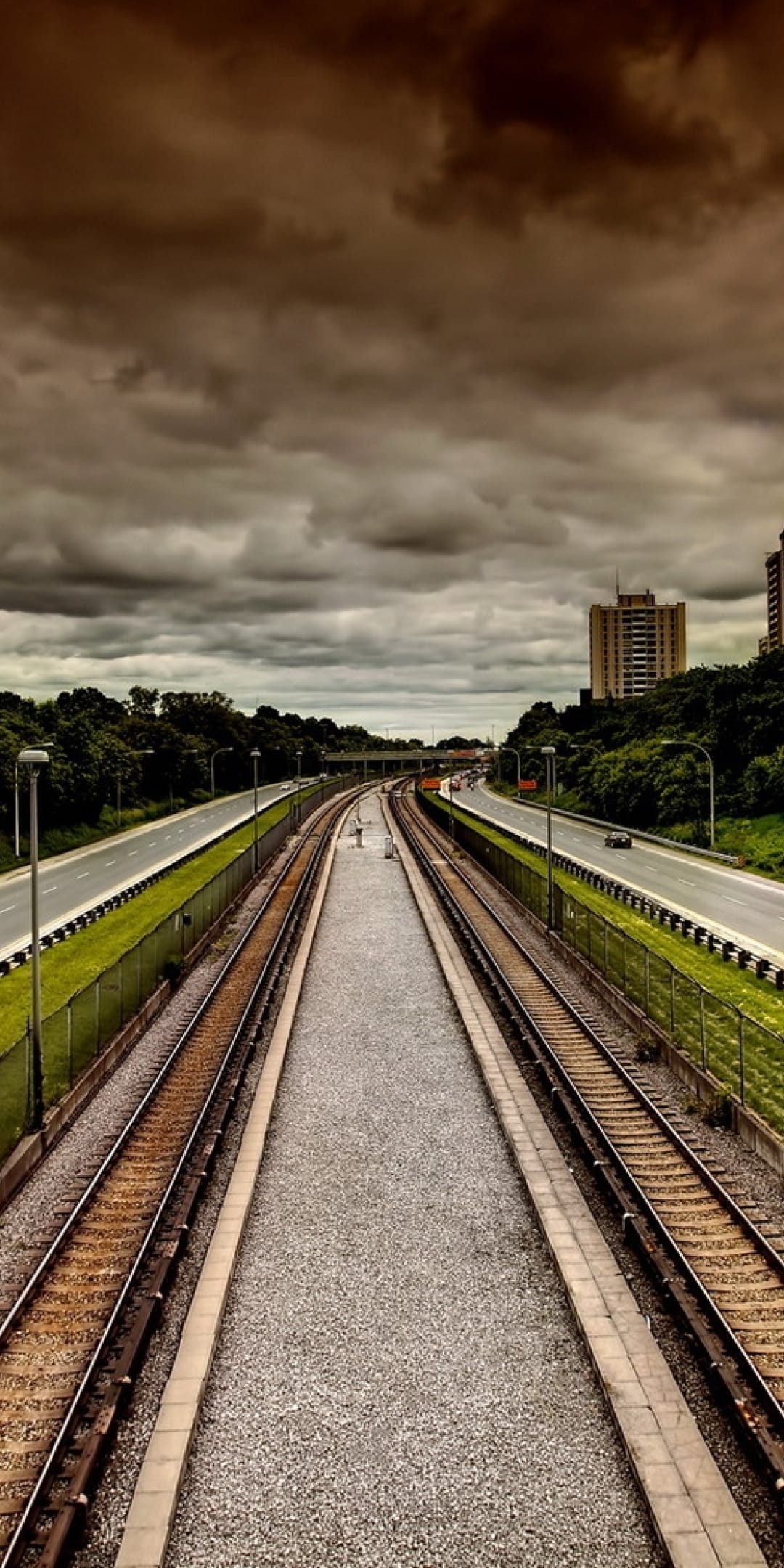 Railway tracks lead toward a hazy city skyline under heavy gray clouds flanked by green grass and highways.