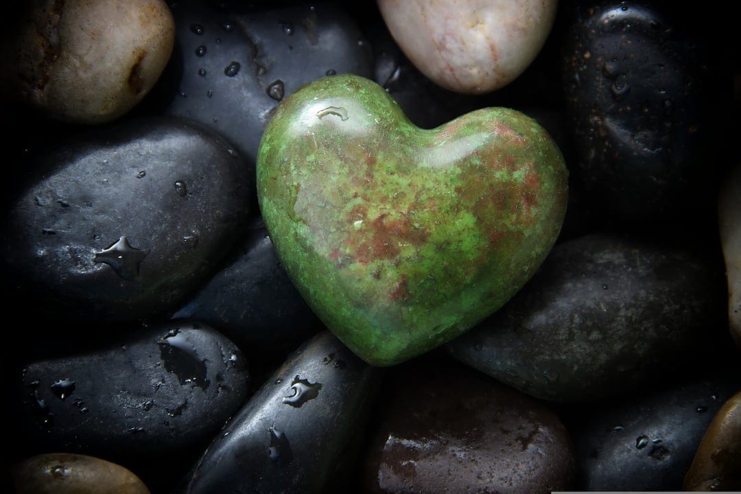 Green heart-shaped stone resting on damp black river rocks with visible water droplets and smooth textures.