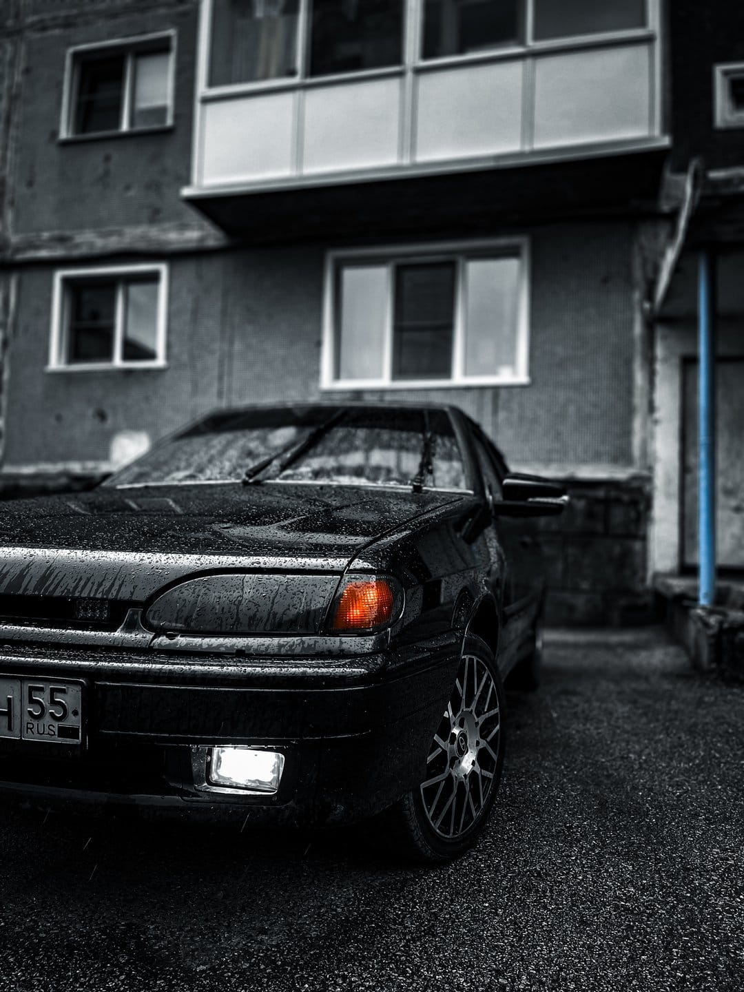 Black sedan with glowing headlights parked on wet asphalt near a grey apartment building on a rainy urban night.