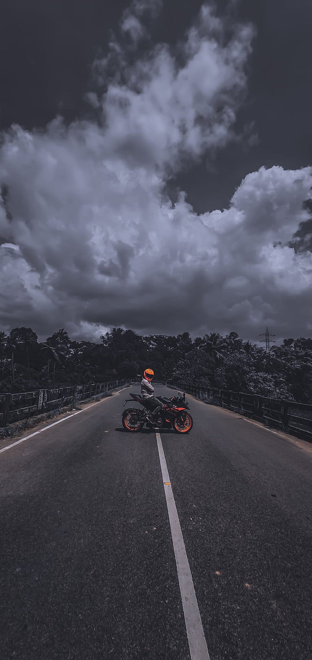 Motorcyclist in a yellow helmet on an orange bike pauses on a bridge road under a dark, dramatic stormy sky.
