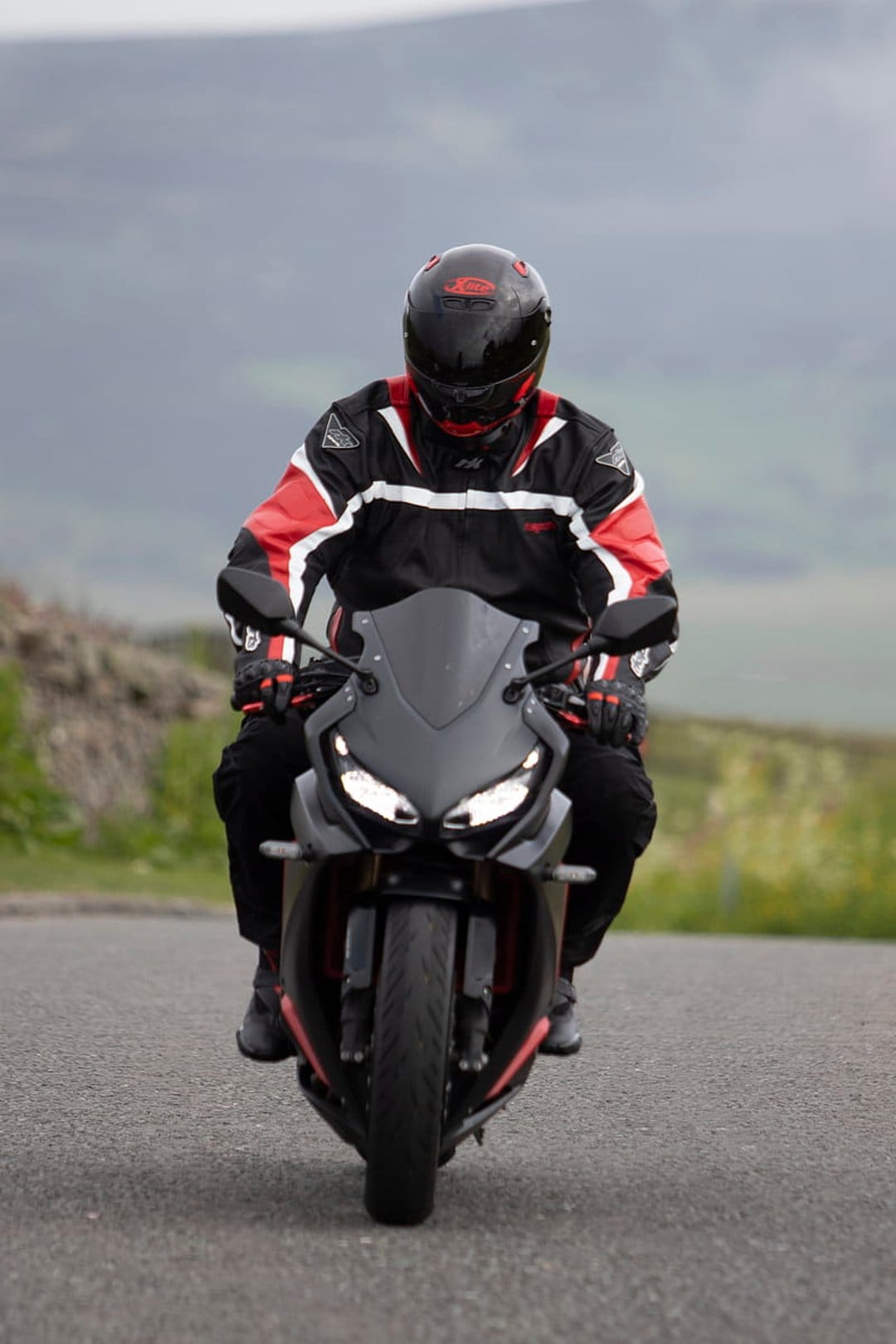Motorcyclist in red and black gear leans a sportbike on a winding asphalt road with blurred hills in the distance.