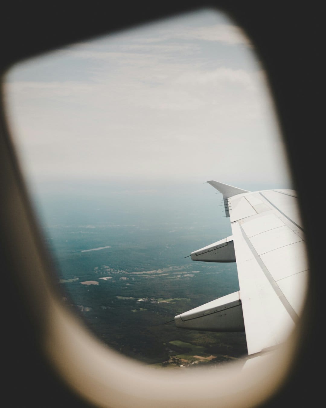 Airplane wing extending over a vast green valley with small buildings under a hazy blue sky from a window view.