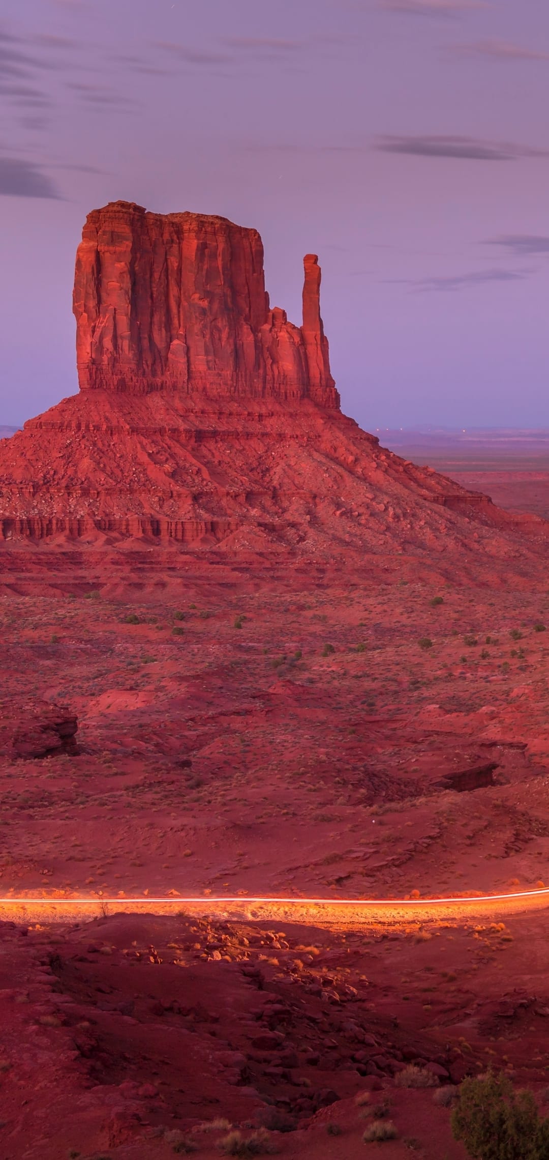 Monument Valley buttes rise against a purple twilight sky with glowing vehicle light trails on a desert road.