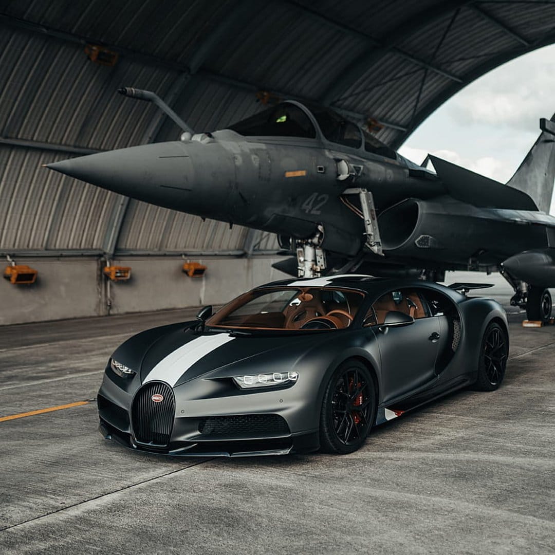 Bugatti Chiron hypercar with a white stripe parked beneath a silver fighter jet inside a dimly lit industrial hangar.