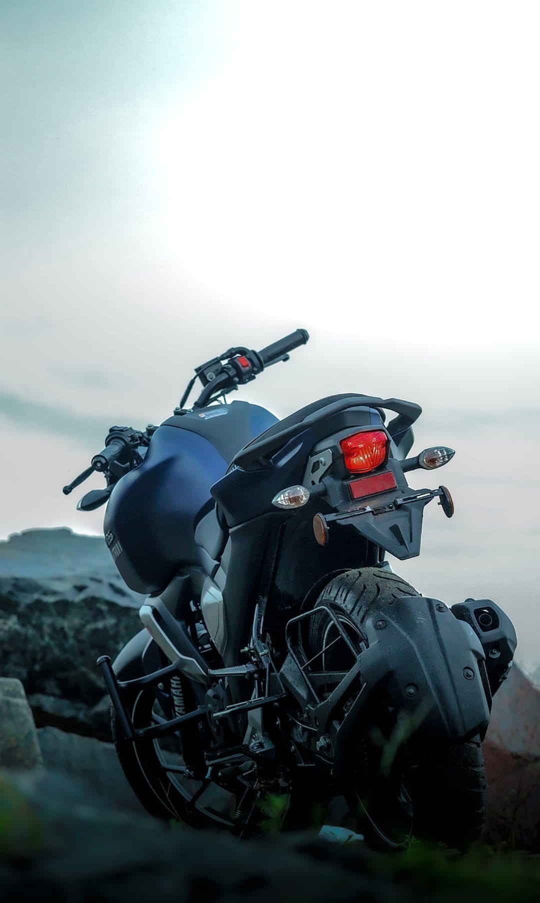 Yamaha FZ motorcycle rear view with a glowing red taillight parked on jagged rocks under a hazy twilight sky.