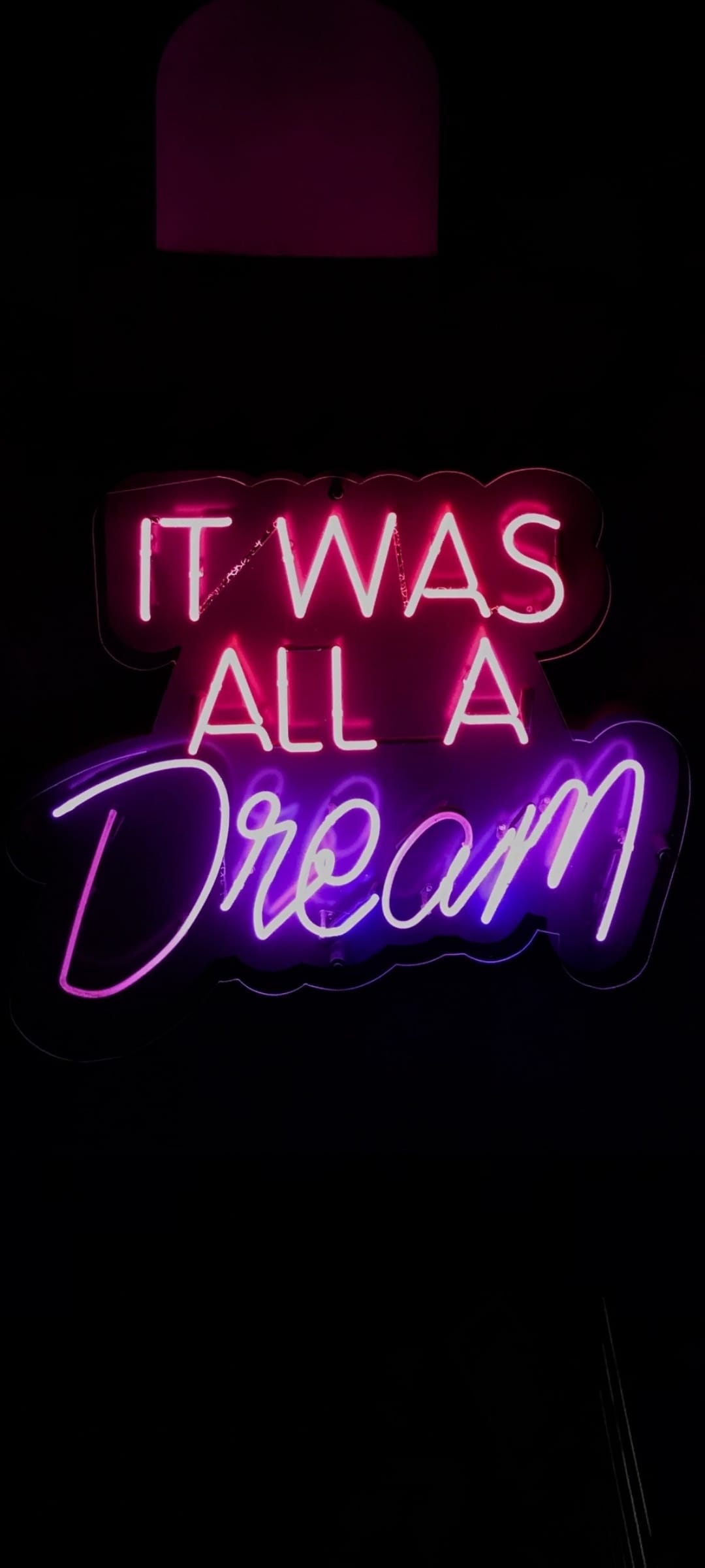 Neon sign reading "It Was All A Dream" glows in hot pink and purple hues against a dark, shadowy background.