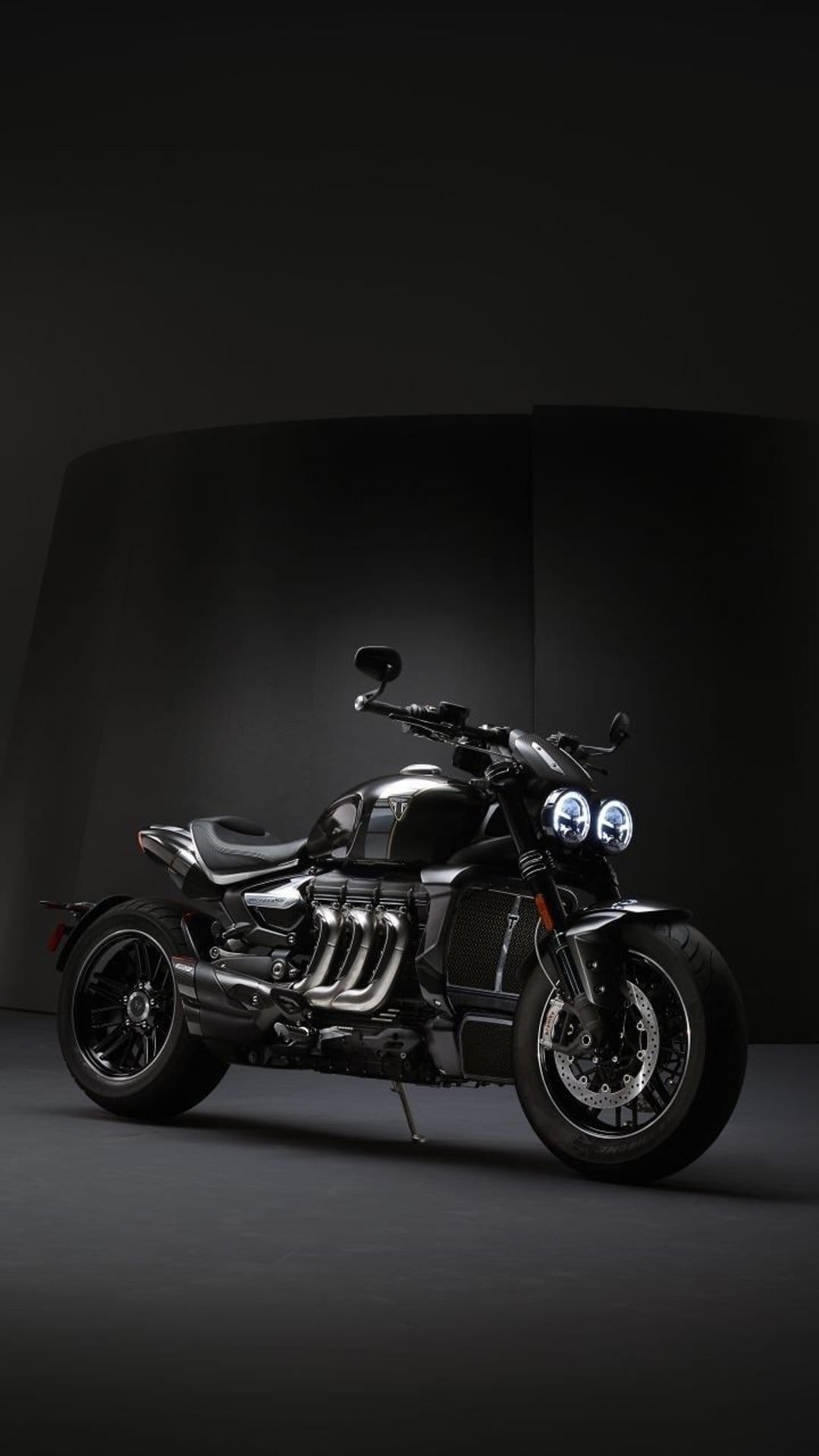 Black Triumph Rocket 3 motorcycle with a large engine displayed in a dark studio under soft spotlighting.