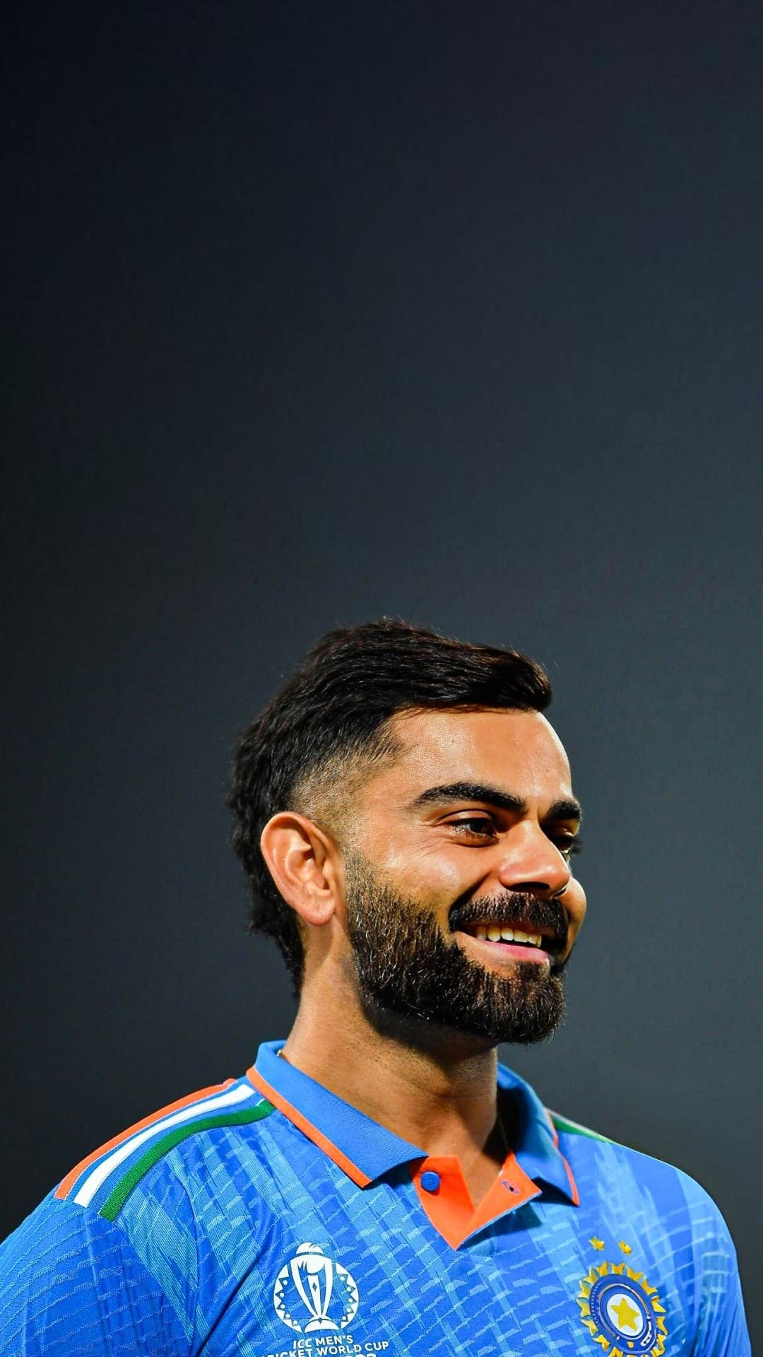 Virat Kohli smiles in a blue Indian cricket jersey with tricolor shoulder accents and World Cup branding.