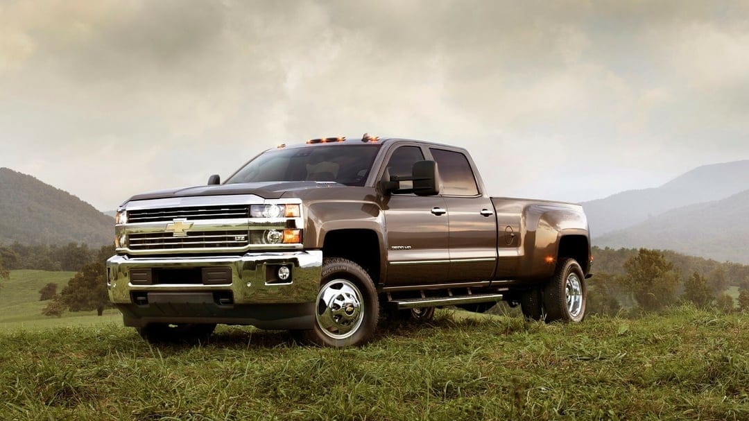 Bronze Chevrolet Silverado 3500 HD dually truck parked on a green grassy hill against a misty mountain range.