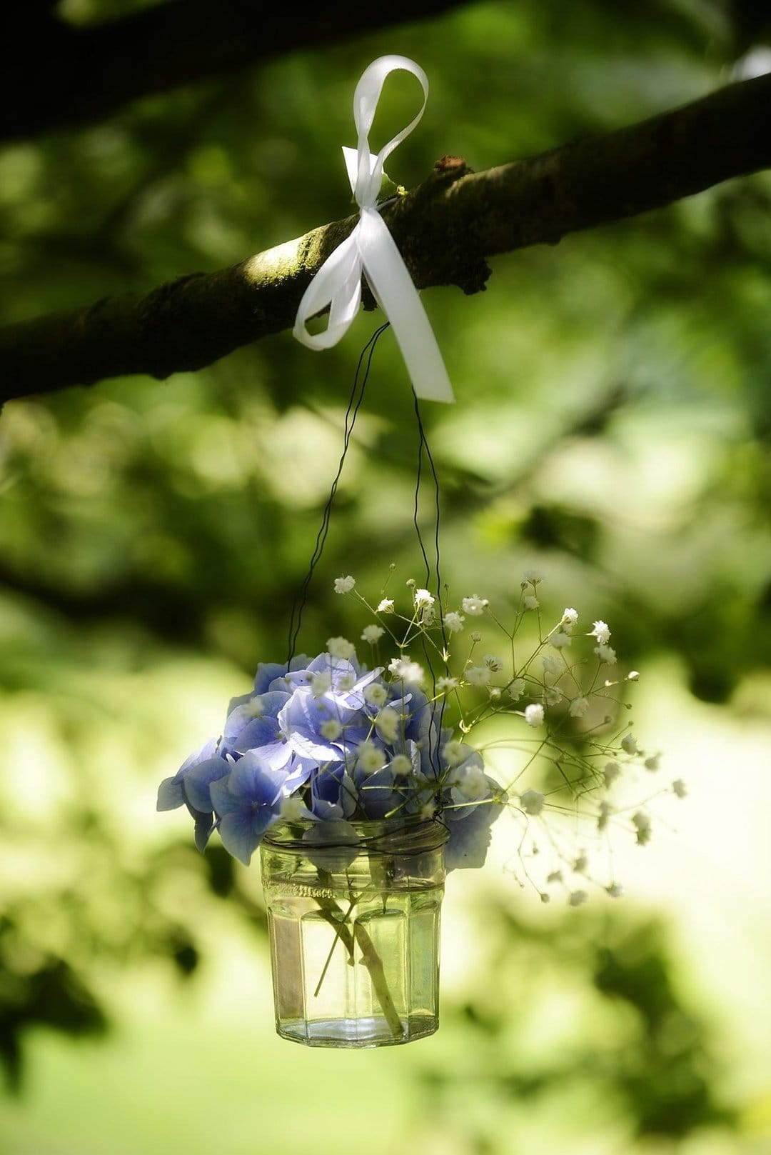 Blue hydrangea bouquet and baby's breath in a glass jar tied to a tree branch with a white ribbon.