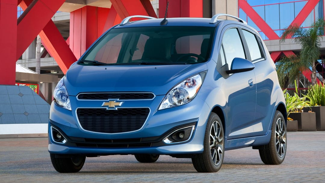 Blue Chevrolet Spark parked in front of red structural beams and palm trees under bright daylight.