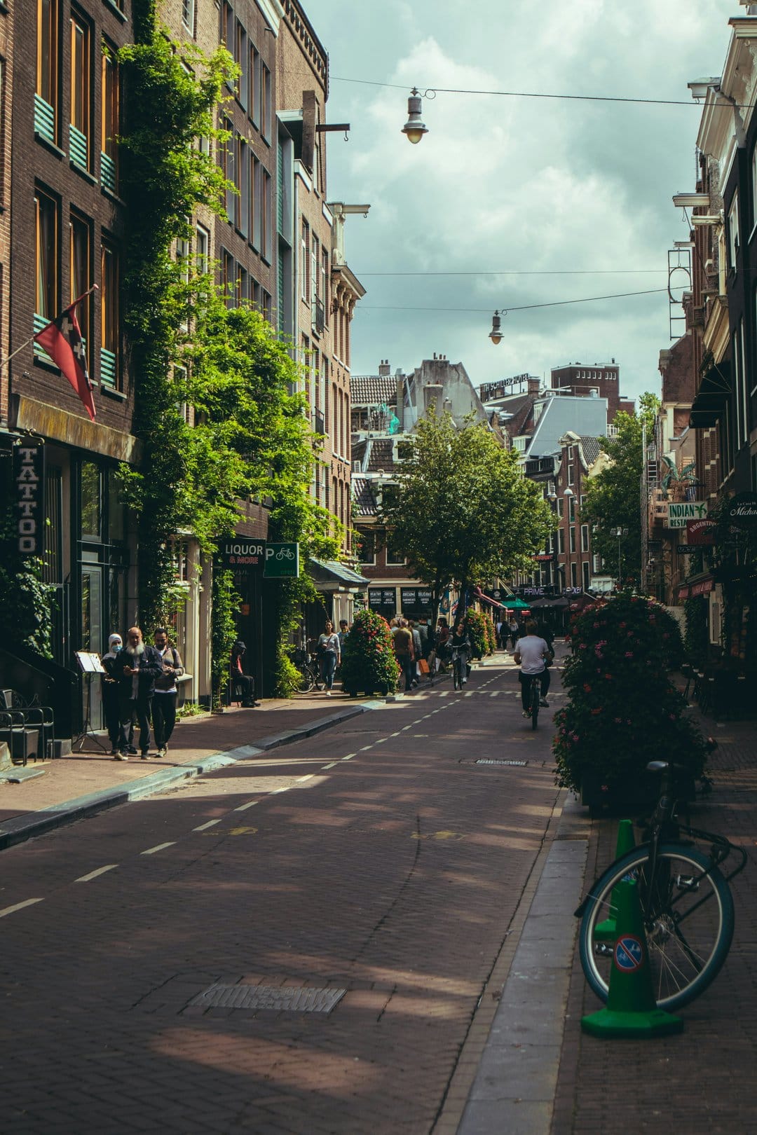 Cyclists ride past ivy-covered brick houses on a cobblestone Amsterdam street under soft morning sunlight.