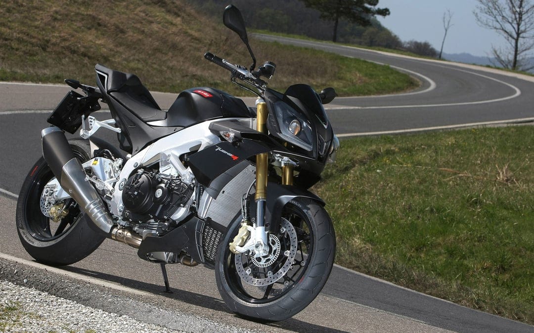 Black and silver Aprilia Tuono V4 motorcycle parked on a winding asphalt road with rolling green hills.