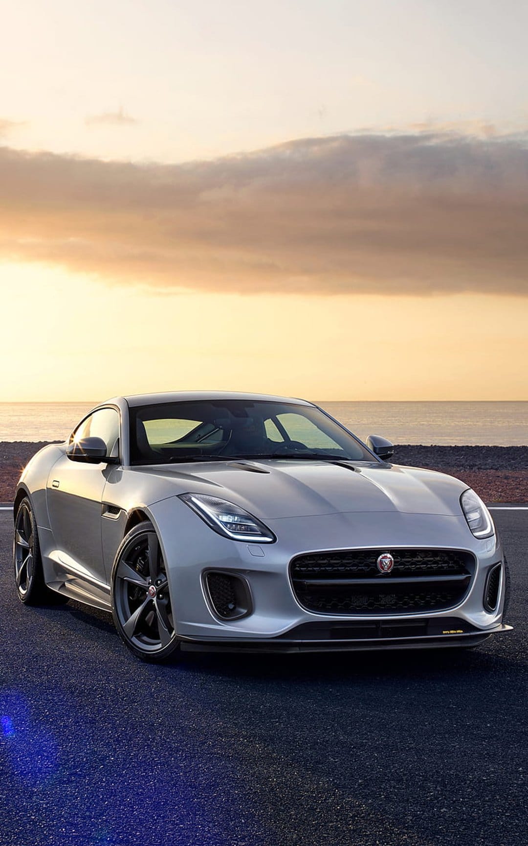 Silver Jaguar F-Type Coupe parked on a seaside road during a golden sunset with orange clouds and ocean waves.