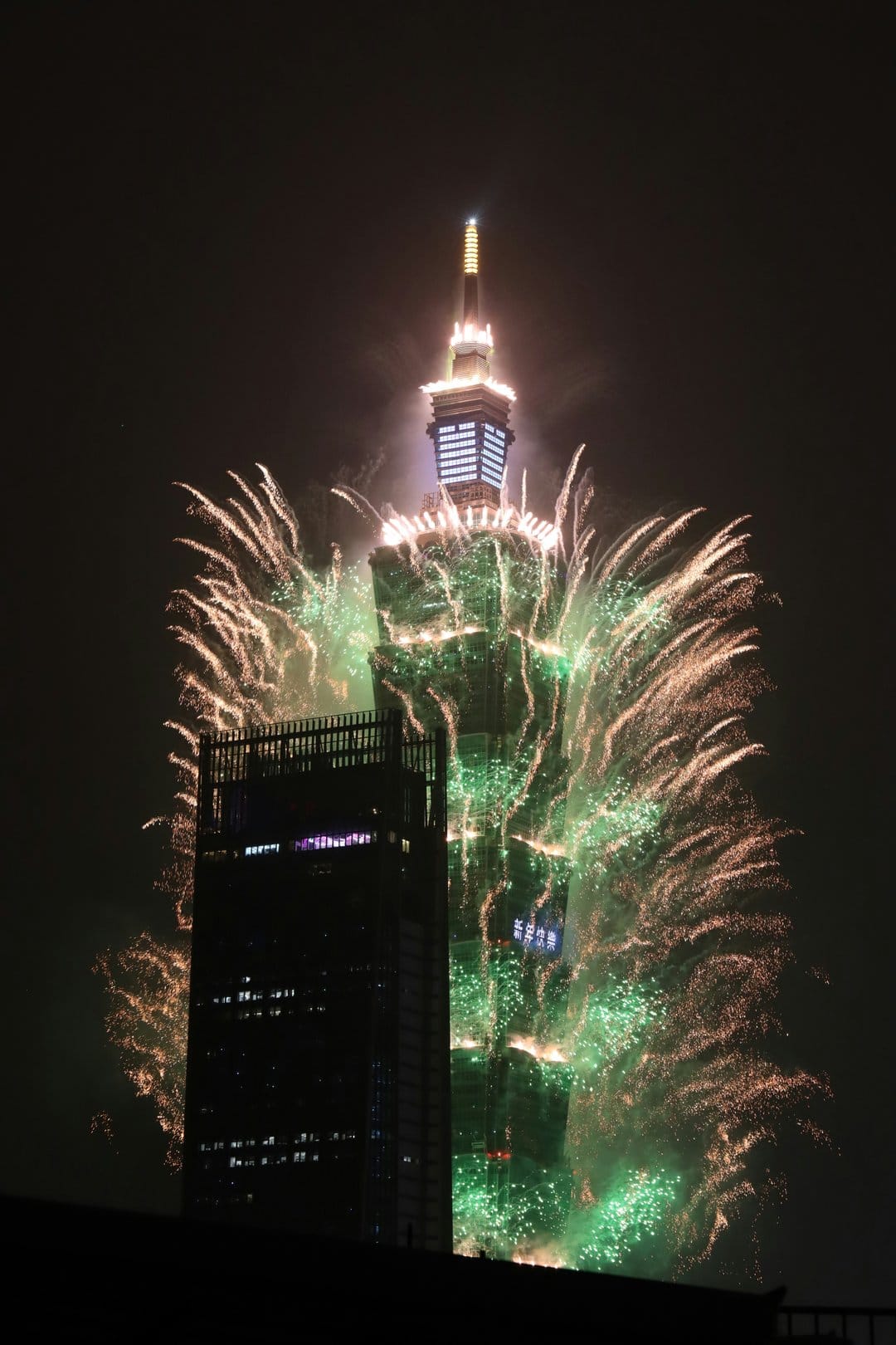 Taipei 101 skyscraper glows with green lights as vibrant gold and red fireworks explode in the dark night sky.