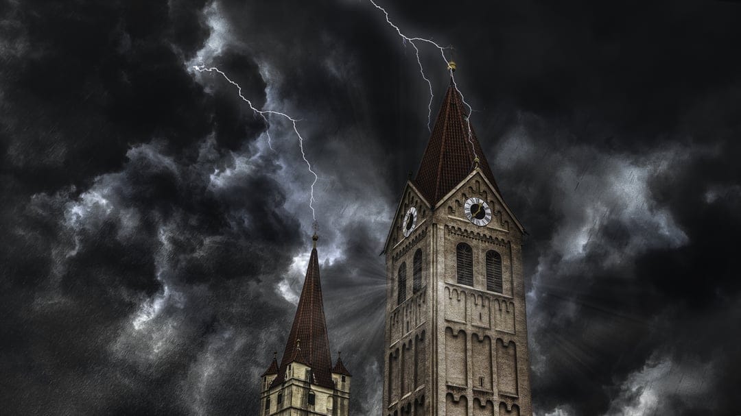 Two stone church spires with a clock face are illuminated by a bright lightning strike under a dark stormy sky.