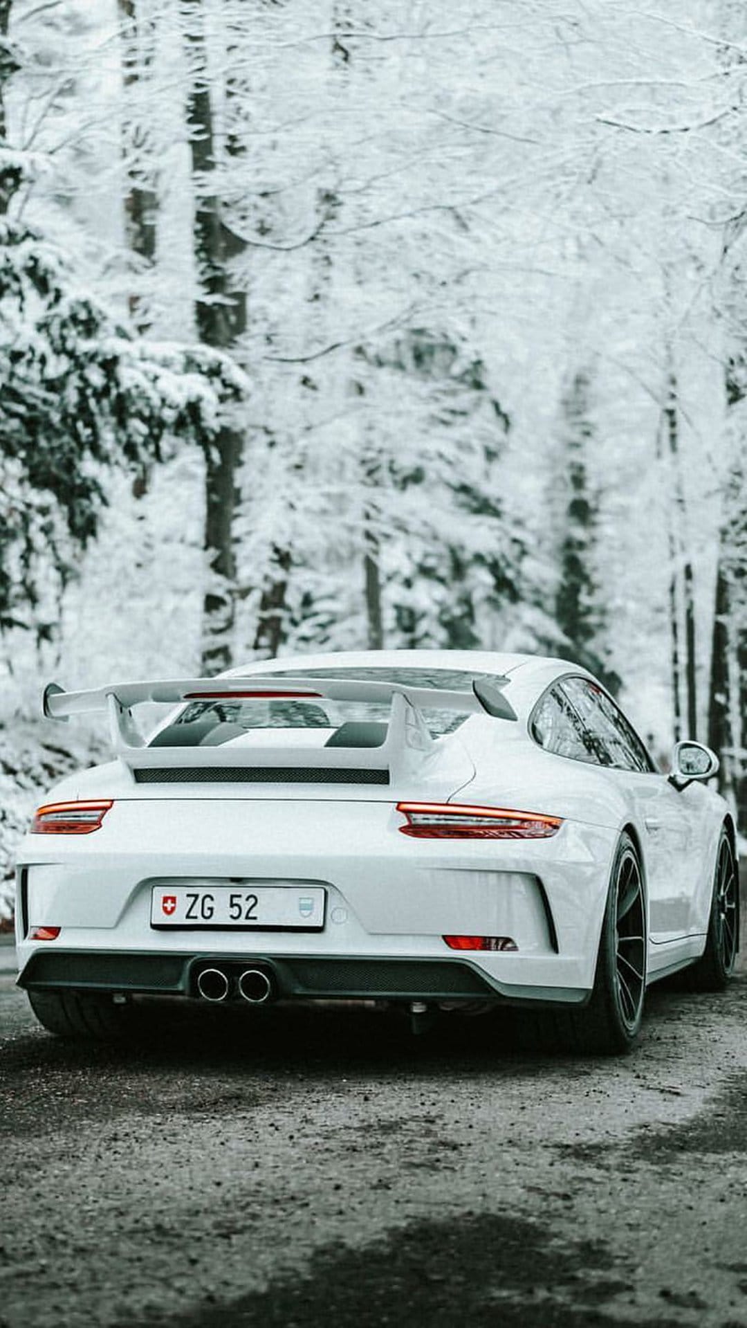 White Porsche 911 with a large rear wing parked on a wet forest road surrounded by snow-covered pine trees.