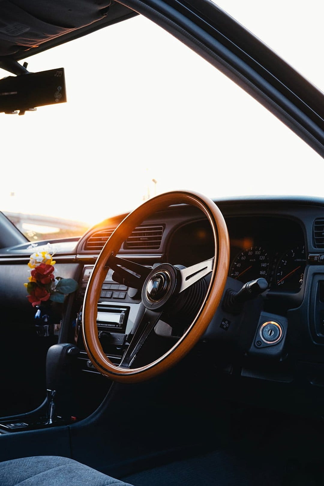 Wooden steering wheel and vintage car dashboard captured in golden sunlight with flowers on the passenger seat.