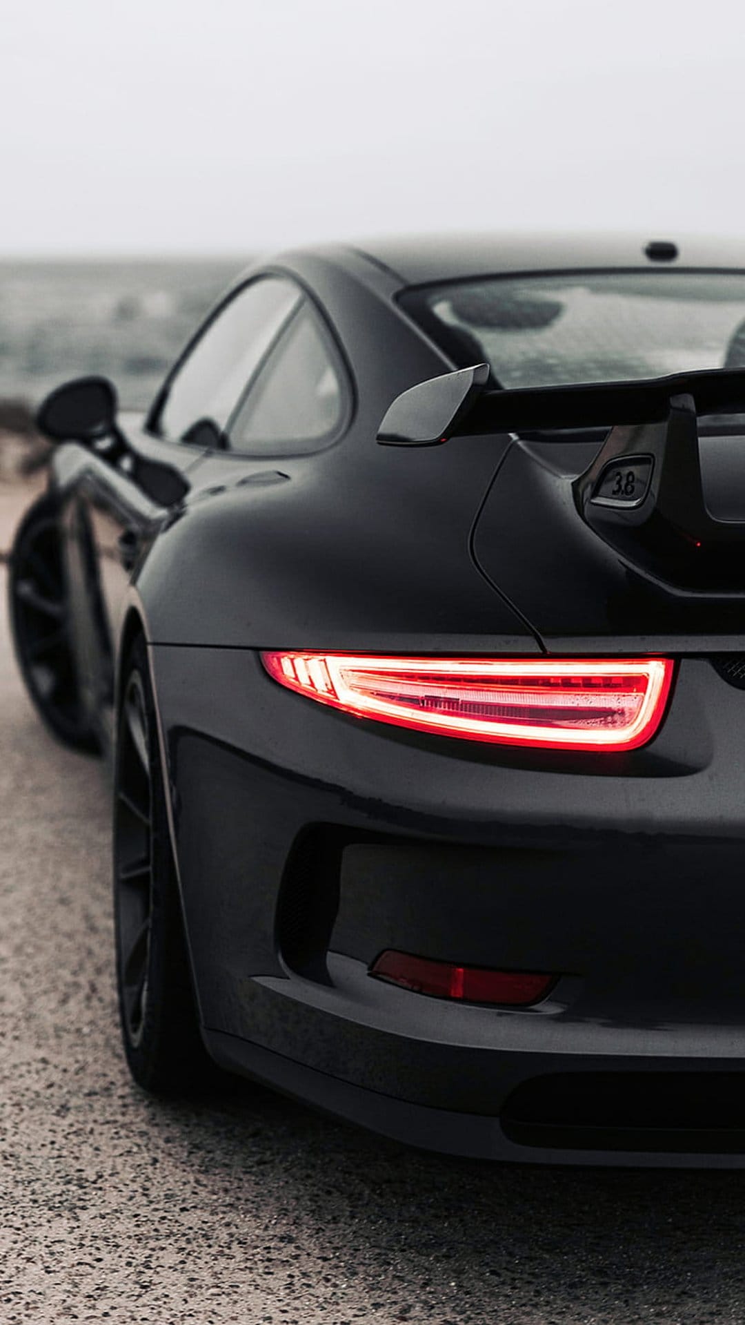 Porsche 911 GT3 rear profile with glowing red taillights and wing on a gravel coastal road at twilight.